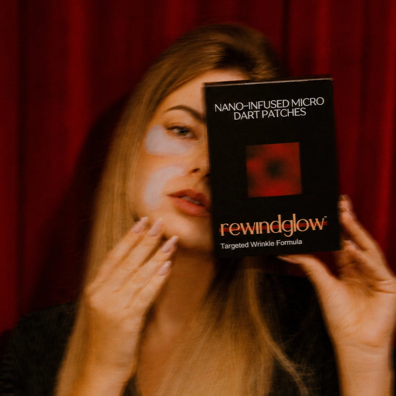 A woman holds a black box of Rewindglow Nano-Infused Micro Dart Patches in front of her face, with a red curtain background.